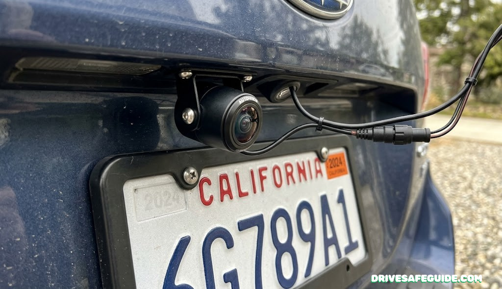 Close-up of an aftermarket backup camera mounted above a license plate, showing the lens and wiring connection